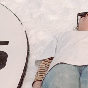 A Person Wearing Sunglasses Lying on the Snow Beside a Surfboard