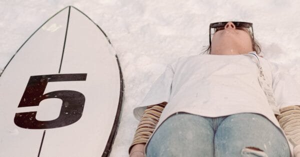 A Person Wearing Sunglasses Lying on the Snow Beside a Surfboard
